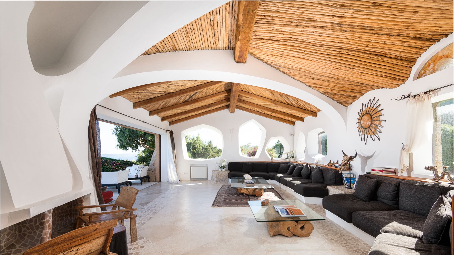 a large living room of a villa in Costa Smeralda, with reeds decorating the ceiling, a very long dark sofas and white walls with big curved windows